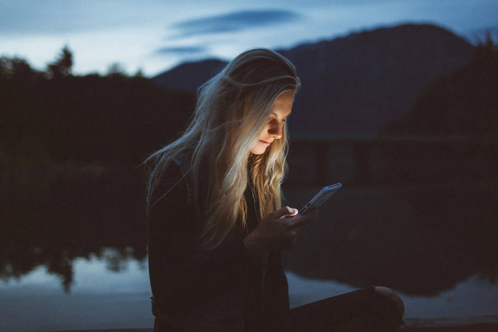 A woman checking her phone at dusk beside a lake